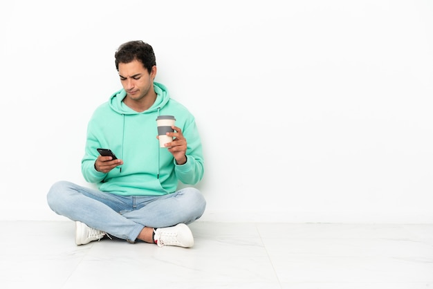 Caucasian handsome man sitting on the floor holding coffee to take away and a mobile