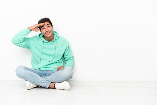 Caucasian handsome man sitting on the floor looking far away with hand to look something