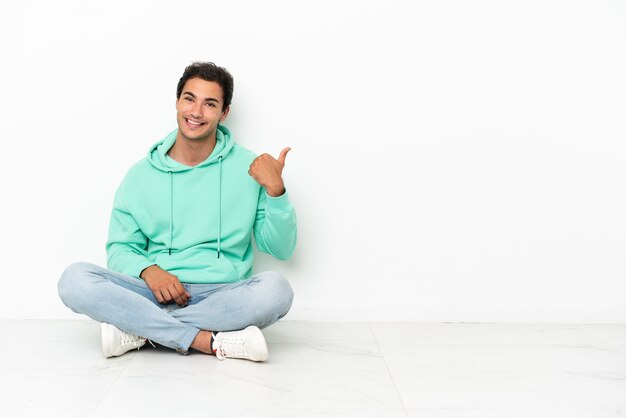 Caucasian handsome man sitting on the floor pointing to the side to present a product