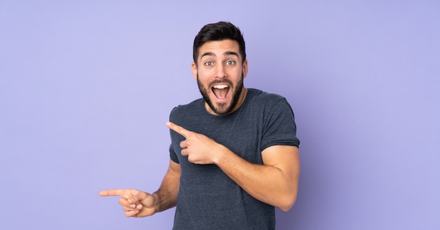 Caucasian handsome man surprised and pointing side over purple wall