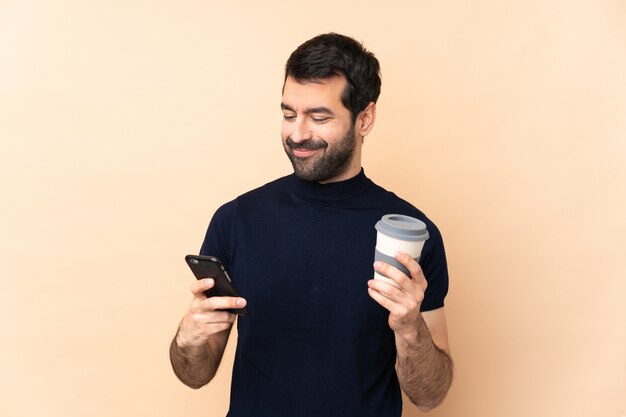 Caucasian handsome man over wall holding coffee to take away and a mobile