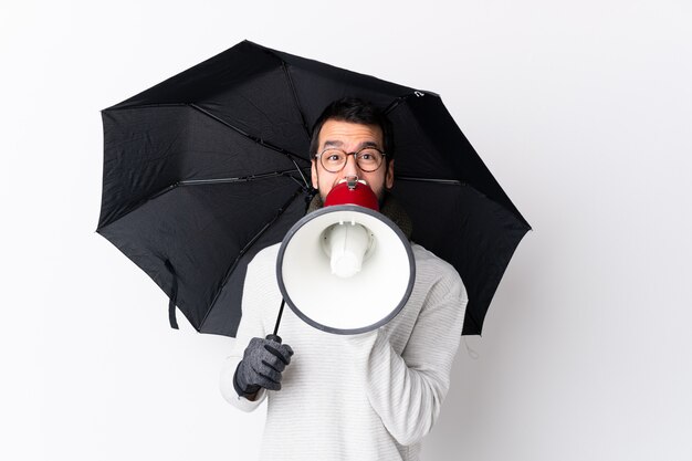 Caucasian handsome man with beard holding an umbrella over white wall shouting through a megaphone