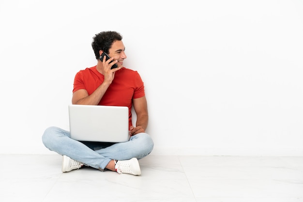 Un bell'uomo caucasico con un laptop seduto sul pavimento che tiene una conversazione con il telefono cellulare con qualcuno