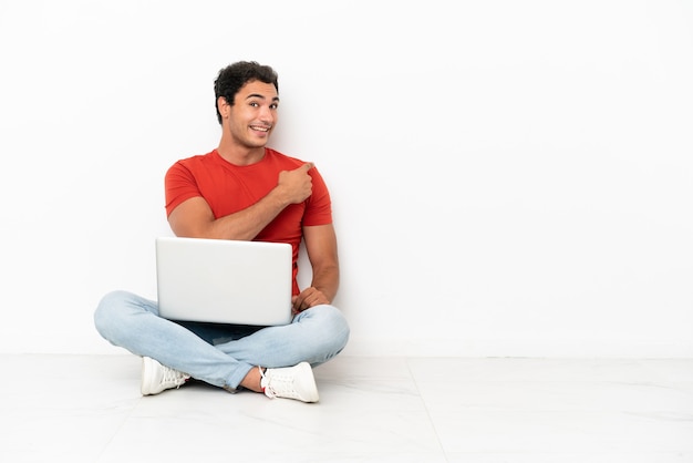 Caucasian handsome man with a laptop sitting on the floor pointing back