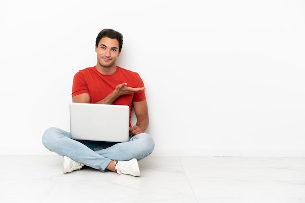 Caucasian handsome man with a laptop sitting on the floor presenting an idea while looking smiling towards