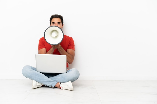 Caucasian handsome man with a laptop sitting on the floor shouting through a megaphone