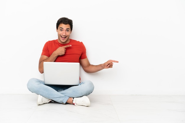 Caucasian handsome man with a laptop sitting on the floor surprised and pointing side