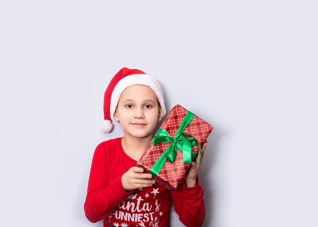 Il bambino caucasico in cappello di babbo natale tiene la confezione regalo su sfondo chiaro spazio di copia per il disegno o il testo banner modello di mockup di natale e capodanno