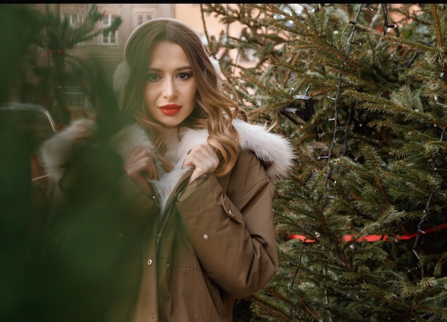 Caucasian lady in a knitted sweater and fluffy headphones. Outdoor portrait of young beautiful happy smiling girl is wearing fur coat near decorated Christmas tree. Poland