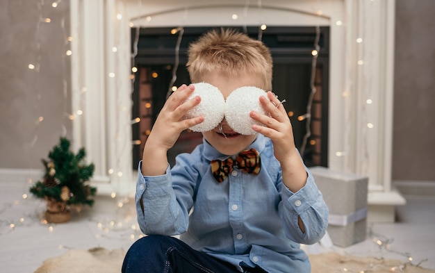 A Caucasian little boy in a blue shirt with a butterfly closed his eyes with Christmas balls