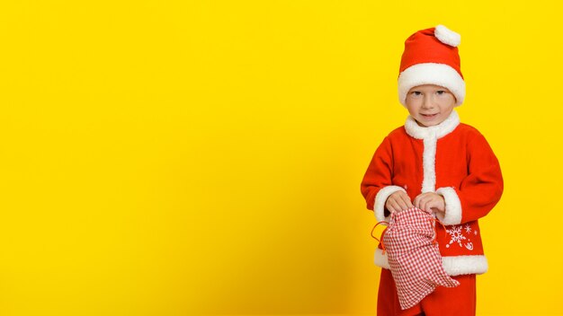 caucasian little boy in a festive Santa Claus costume stands with an open sack of New Years gifts