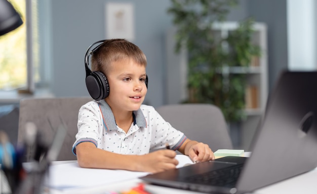 Caucasian little boy in headphones using laptop for online studying Schoolboy sitting at desk and writing in notebook Education at home