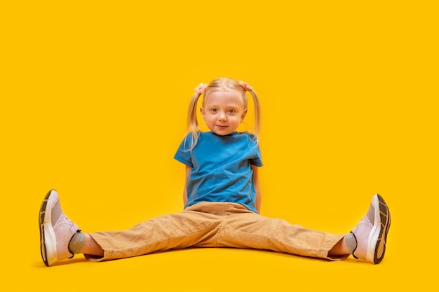 Caucasian little fairhaired sport girl with two ponytails wears blue tshirt and yellow pants sits on floor with legs spread wide