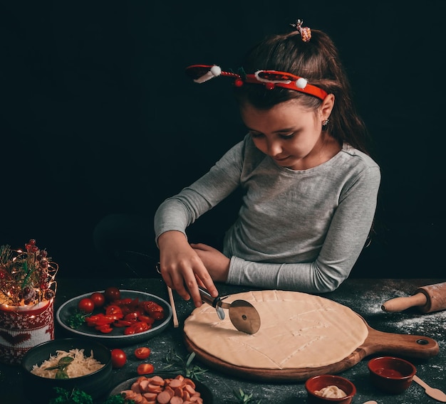 A Caucasian little girl cuts a Christmas tree with a round moon knife on a pizza dough