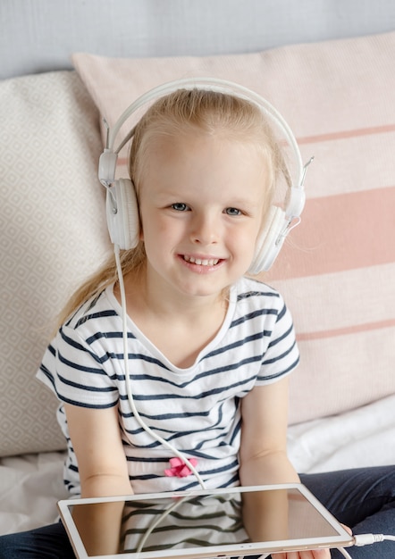 ベッド、家の内部、現代装置技術スカンジナビアスタイルのタブレットを見てヘッドフォンで白人少女