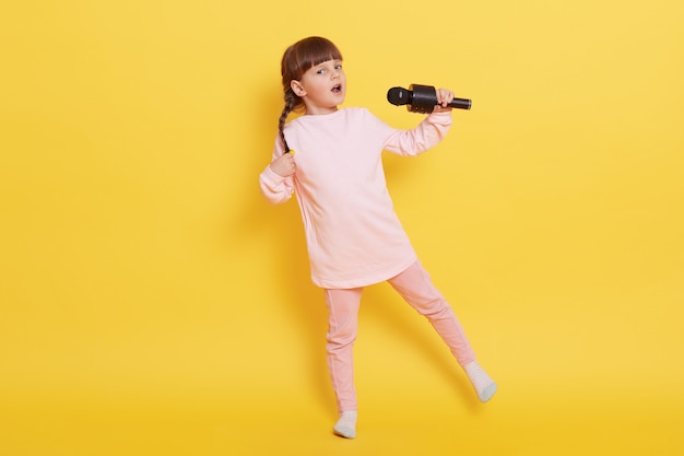 Bambina caucasica che canta con il microfono e balla, piccolo artista sveglio che esegue isolato sopra la parete gialla, abbigliamento casual, cantante affascinante canta per qualcuno.