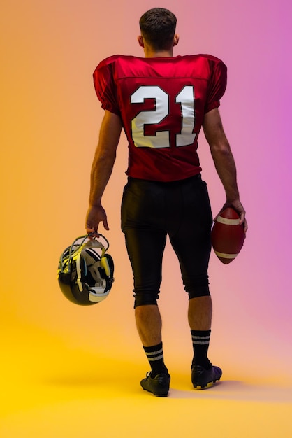 Caucasian male american football player holding helmet and ball with neon yellow and purple lighting