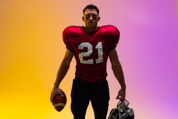 Photo caucasian male american football player holding helmet and ball with neon yellow and purple lighting