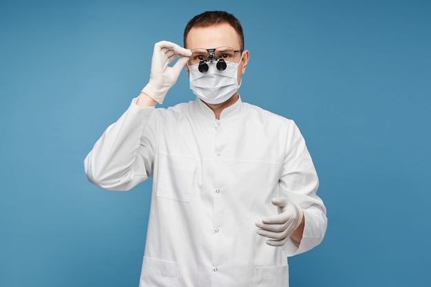 Medico maschio caucasico in una mascherina chirurgica, guanti protettivi e lenti binoculari sullo sfondo blu, isolato con spazio di copia per testo o posizionamento del prodotto