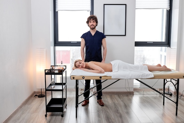 Caucasian male massage therapist stands next to relaxed female client lying on massage table getting massage by him, bearded masseur in blue uniform posing at camera, in spa salon