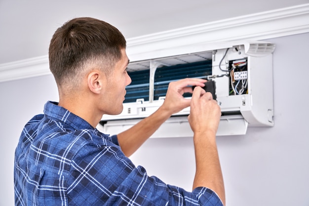 Caucasian male master in blue shirt cleans filters, installs and fixes air conditioner indoors