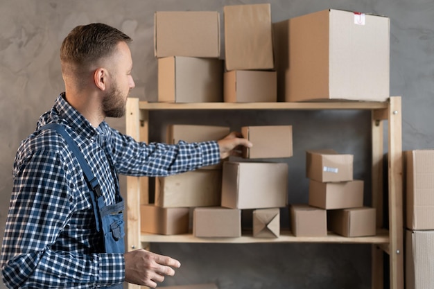 Caucasian male worker dressed in blue overalls stands near a shelf with boxes with his hand laying one of them on a rack