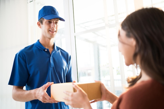 Caucasian man courier in uniform delivering a parcel shipping package to customer Asian woman hands to hand in front of the entrance door at home Home parcel shipping delivery service concept