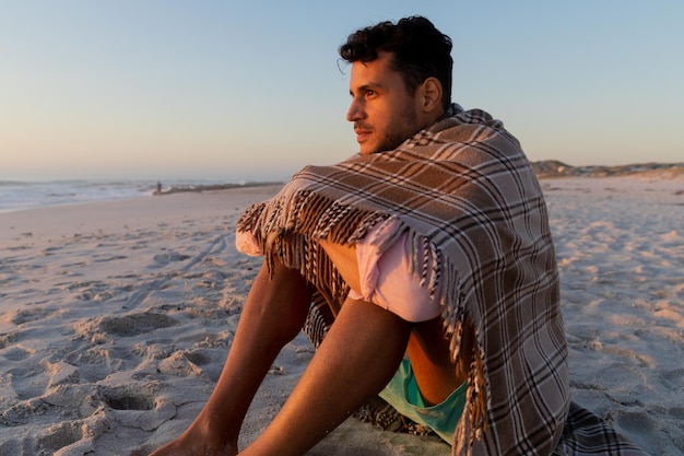 Кавказский мужчина наслаждается временем на пляже, сидя под одеялом, во время заката