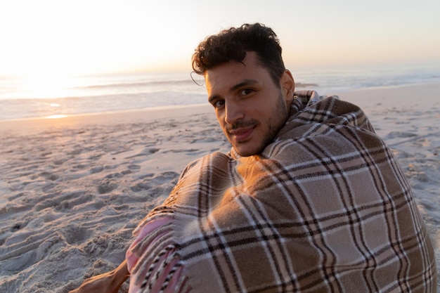 日没時に毛布をかぶって座り、ビーチで時間を楽しむ白人男性
