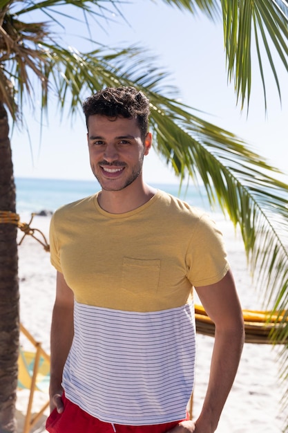 Foto uomo caucasico che si gode il tempo in spiaggia, in piedi accanto a una palma, guardando la telecamera e sorridendo