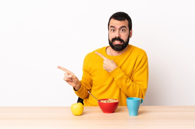 Caucasian man having breakfast in a table frightened and pointing to the side.