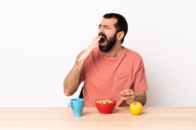 Кавказский человек завтракает в таблице зевая и прикрывая широко открытый рот рукой