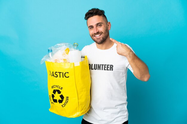 Caucasian man holding a bag full of plastic bottles to recycle isolated on blue background giving a thumbs up gesture