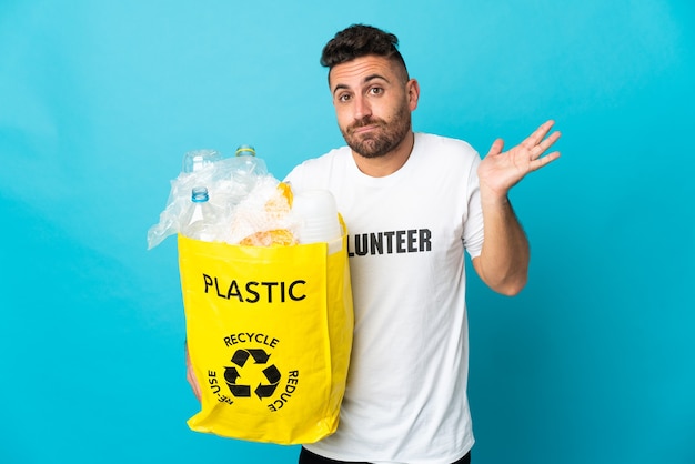 Caucasian man holding a bag full of plastic bottles to recycle isolated on blue background having doubts while raising hands