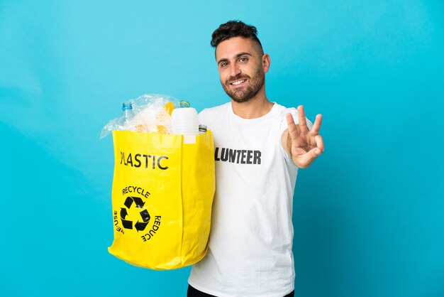 リサイクルするためにペットボトルでいっぱいのバッグを持っている白人男性は、幸せな青で隔離され、指で3つを数えます