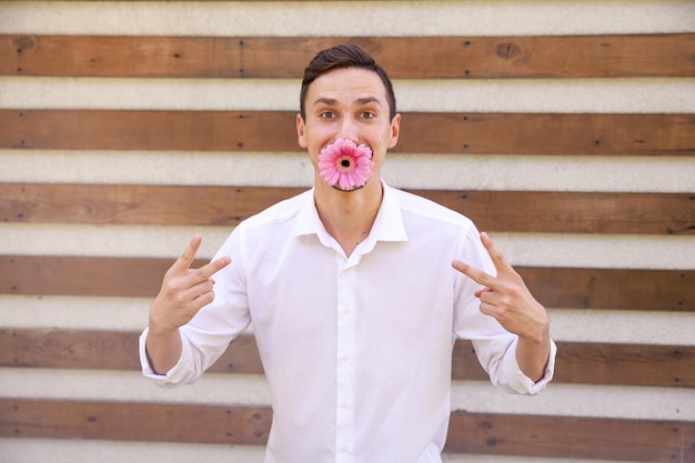 Caucasian man holding a pink flower in his mouth and showing the vsign