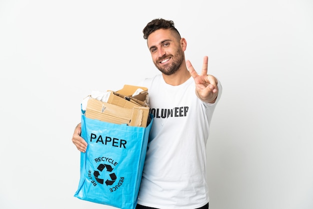 Caucasian man holding a recycling bag full of paper to recycle isolated on white wall smiling and showing victory sign