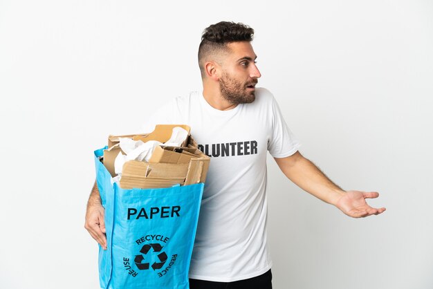 Кавказский мужчина держит мешок для переработки, полный бумаги для переработки, изолирован на белой стене с удивленным выражением лица, глядя в сторону