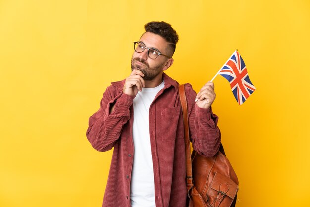 Caucasian man holding an United Kingdom flag isolated on yellow wall and looking up