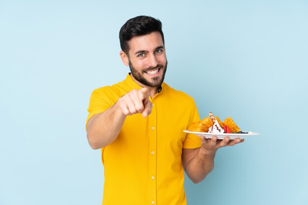 Caucasian man holding waffles on blue wall points finger at you with a confident expression