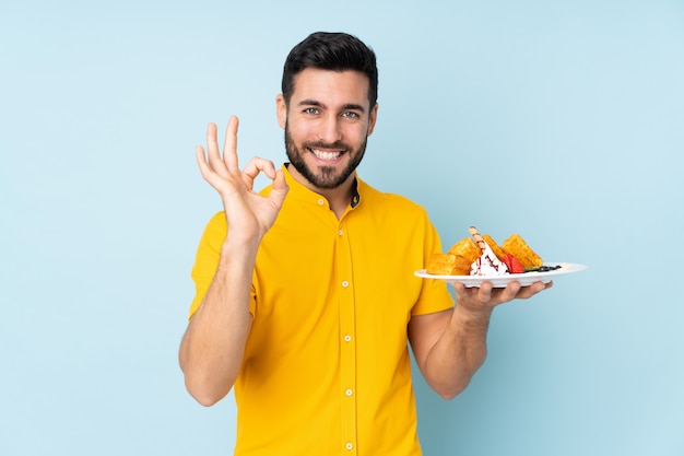 Caucasian man holding waffles on blue wall showing ok sign with fingers