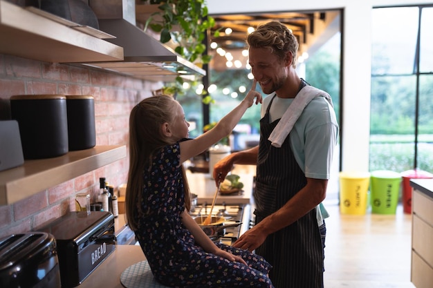 Caucasian man at home, standing by a table, with his daughter touching his nose, cooking pasta together, social distancing and self isolation in quarantine lockdown during coronavirus covid19 epidemic