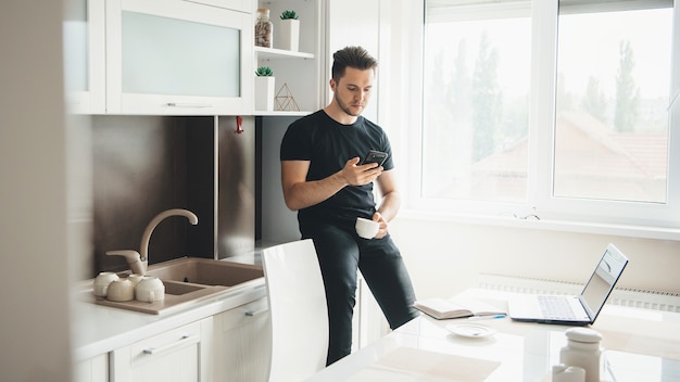 Caucasian man is drinking a coffee and chatting on mobile at home while working remotely on laptop