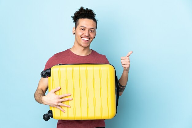 Caucasian man over isolated blue wall in vacation with travel suitcase and with thumb up
