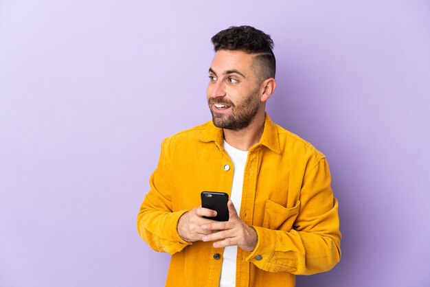 Caucasian man isolated on purple using mobile phone and looking up