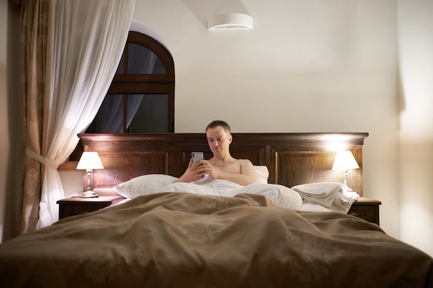 Caucasian man laying in bed in hoter room and chatting by smartphone