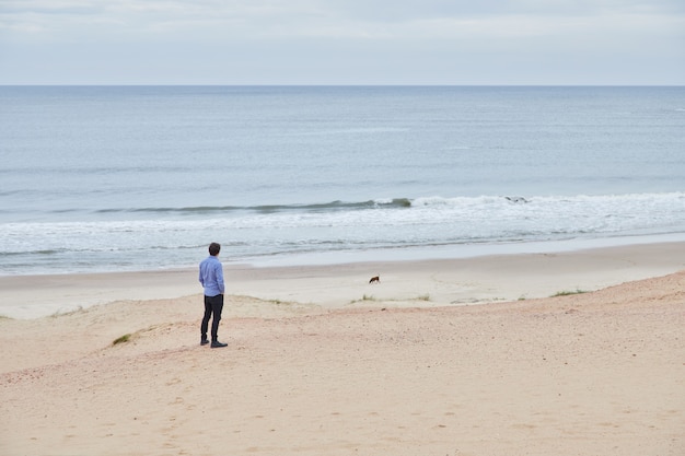 Caucasian man in light blue shirt on the beach looking at the horizon.