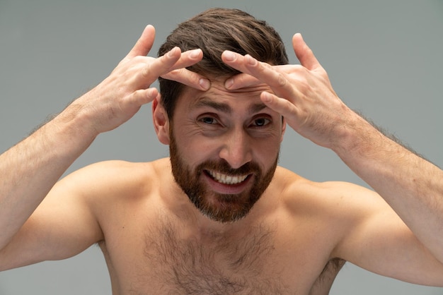 Кавказский мужчина играет со своим лицом раздавить прыщи, изолированные на пастельно-сером фоне студийный портрет концепция косметических процедур по уходу за кожей