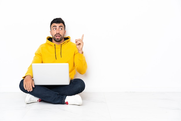 Кавказский мужчина сидит на полу со своим ноутбуком, думая об идее, указывая пальцем вверх