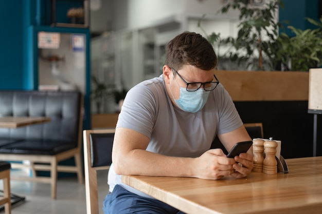 Caucasian man sitting in restaurant and using phone in medical face mask. New Normal concept